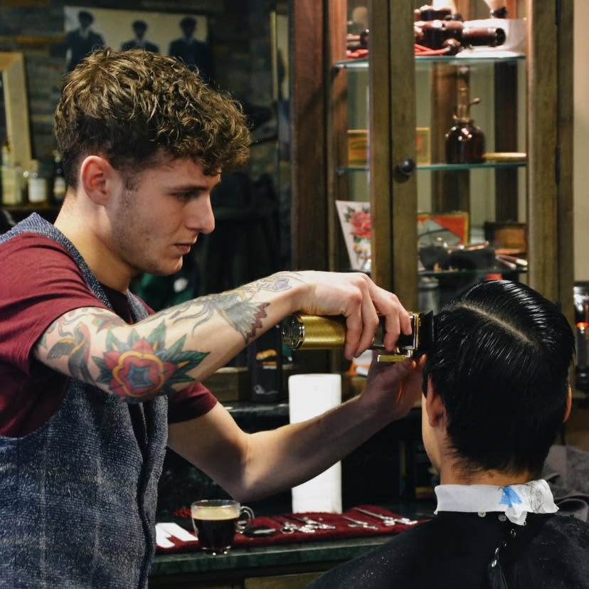 Barbier met getatoeëerde armen werkt een fade bij met de tondeuse, op de voorgrond een glas koffie