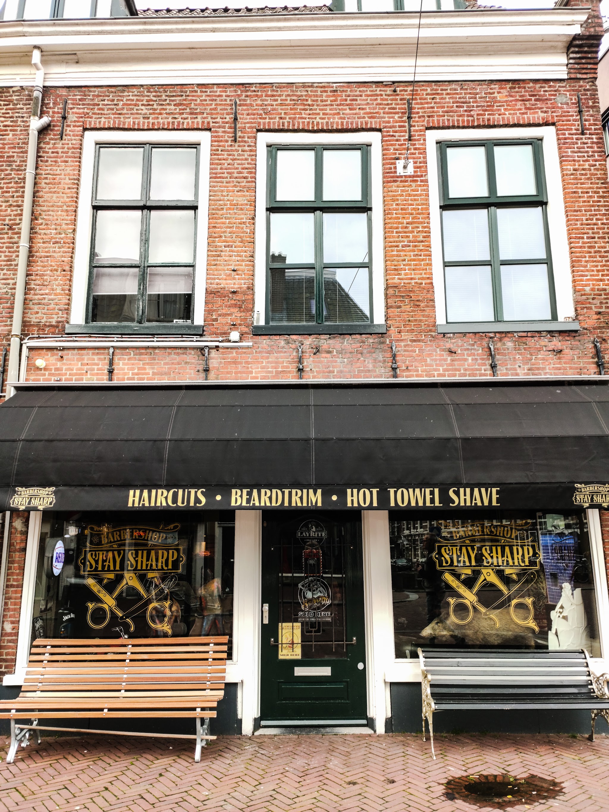 De winkelpui van Barbershop Stay Sharp: zwarte luifel met Haircuts, Beardtrim, Hot towel shave, het gouden logo op beide ramen en twee bankjes voor de deur