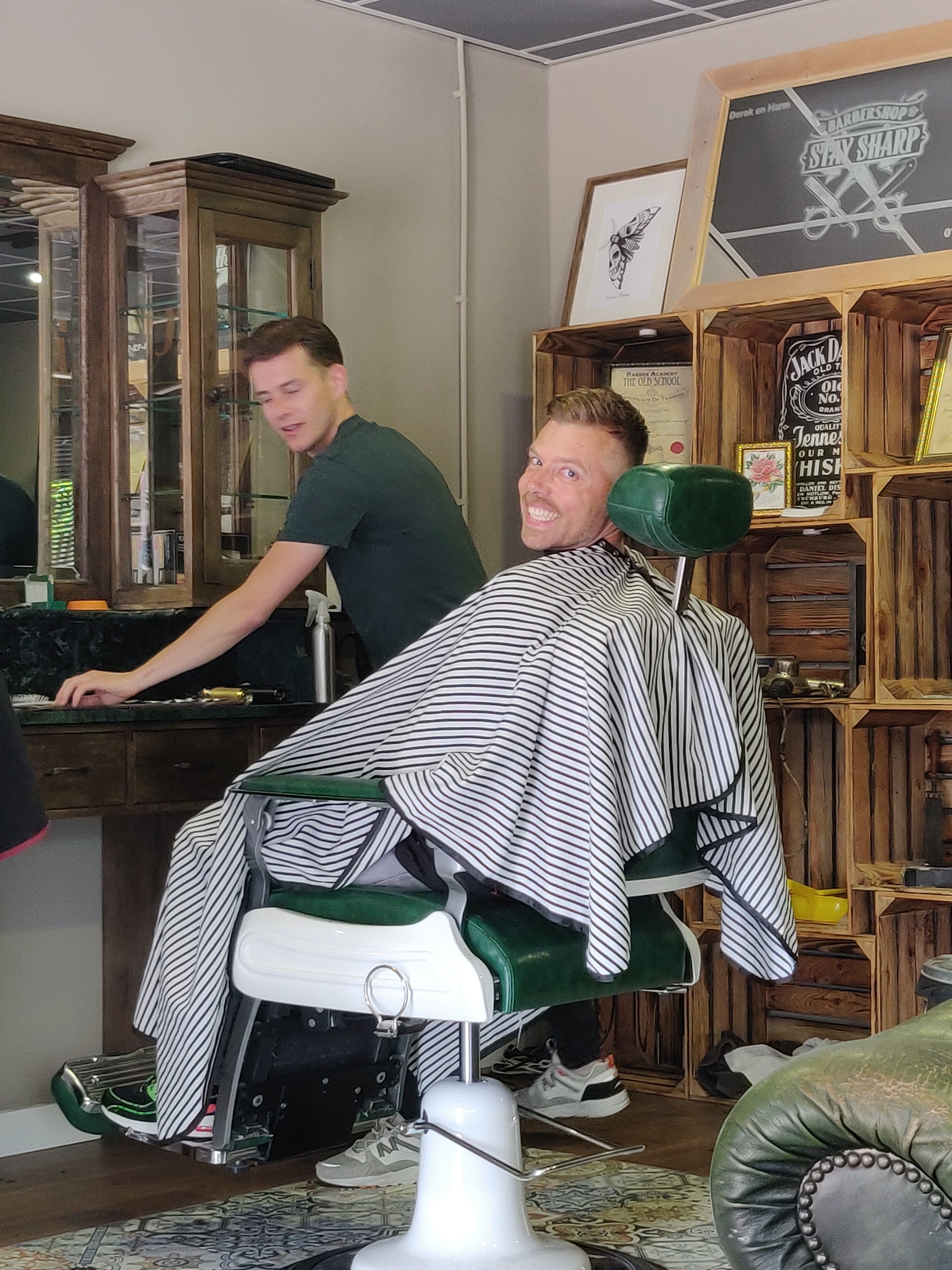 Een barbier aan het werk bij een lachende klant onder een gestreepte kapmantel, met de kratten en het krijtbord met het logo op de achtergrond